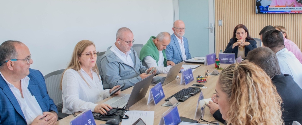 Varias personas reunidas alrededor de una mesa, con computadores portátiles, documentos y tarjetas, mientras algunas conversan.