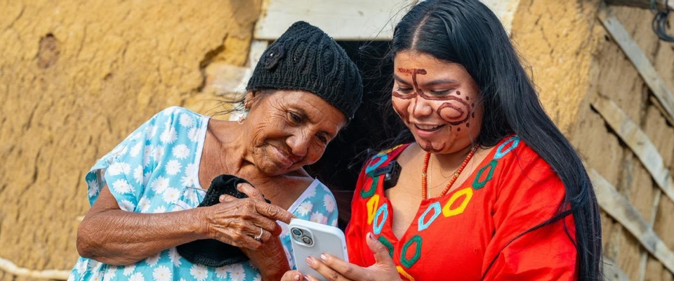 Mujer mayor y joven sonríen mientras miran un celular. Ambas tienen vestimenta colorida; la joven lleva pintura facial.